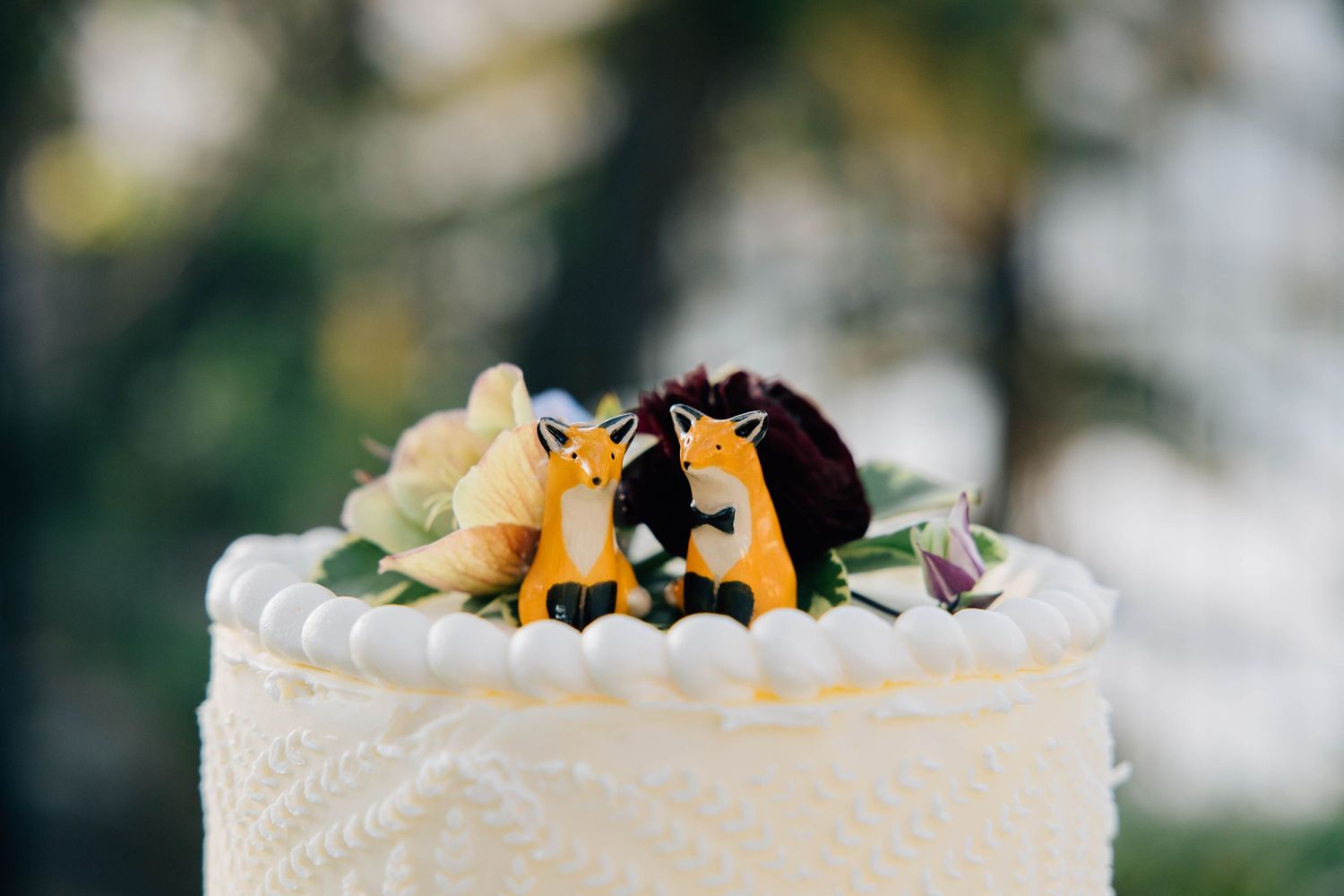 White wedding cake topped with fox bride and groom figurines surrounded by floral decorations.