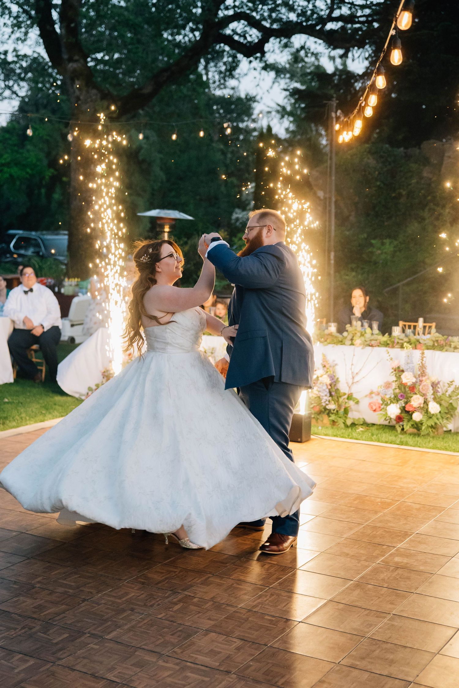 Bride and groom share first dance outdoors at twilight, surrounded by sparkling fountains and warm string lights.