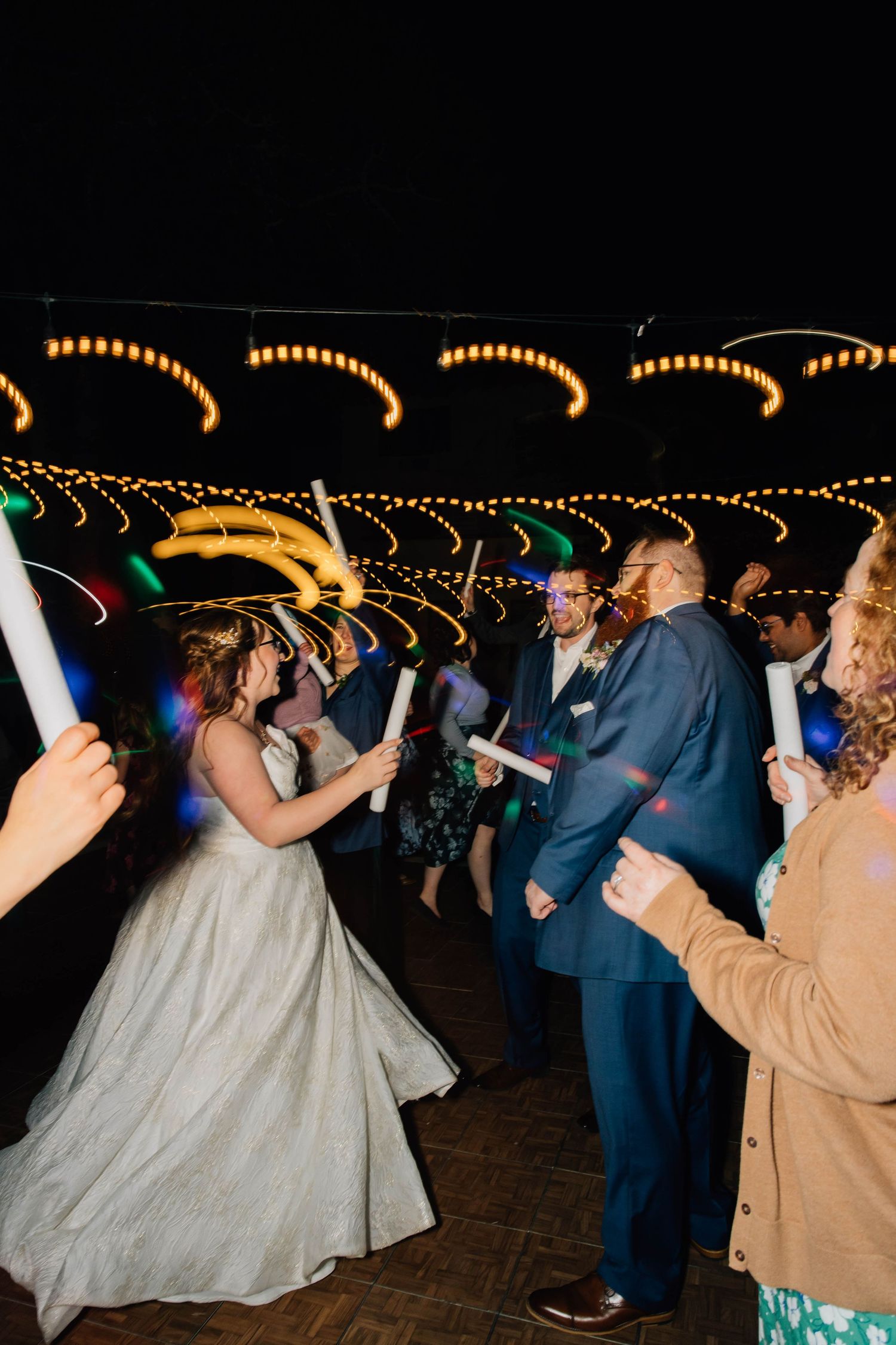 Bride and groom celebrate with glowing light sticks during outdoor wedding reception at night with string lights.