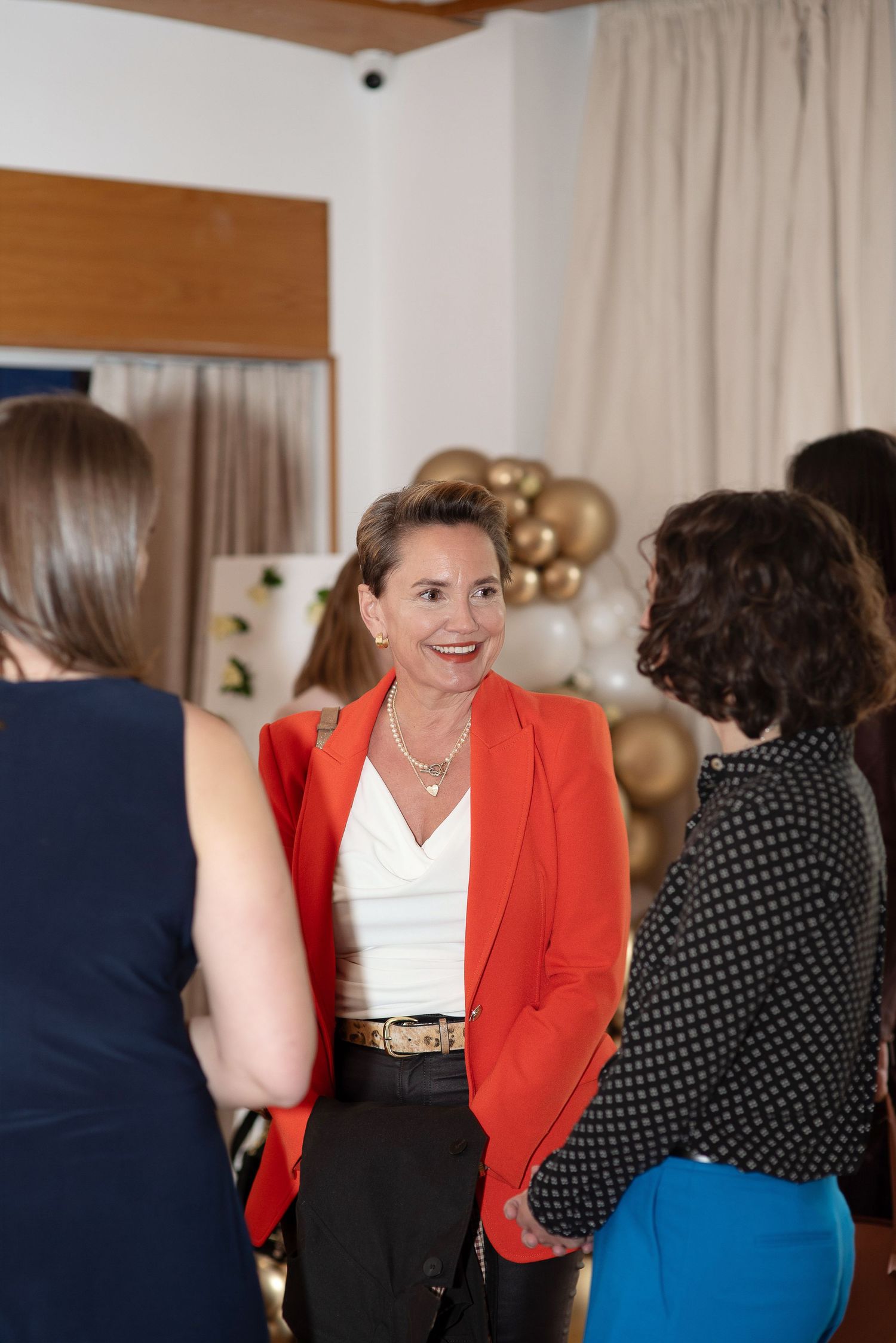Woman in red blazer smiling at a social gathering with gold balloons in the background.