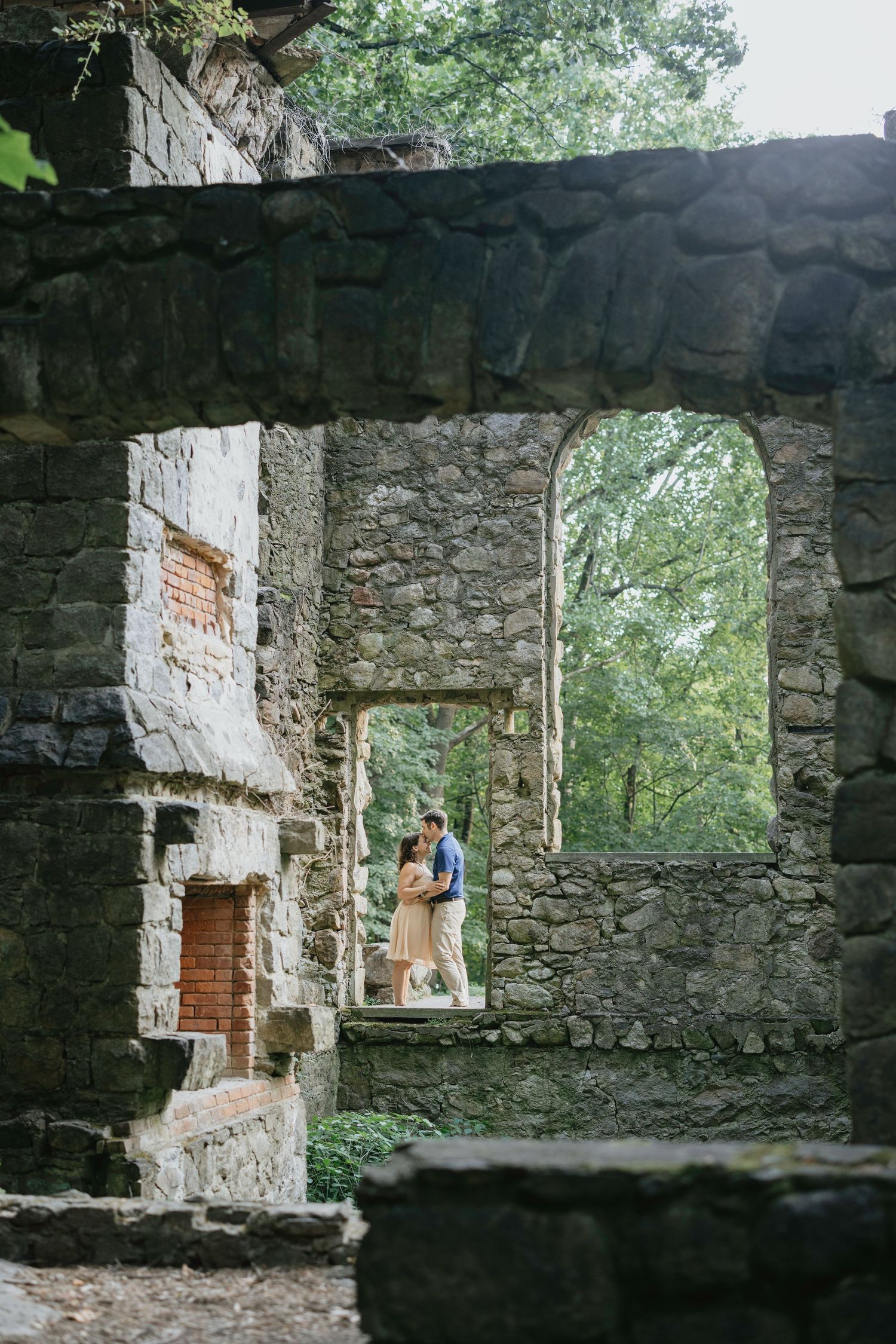 Couple embracing in romantic stone ruins archway surrounded by lush green trees in historic setting.