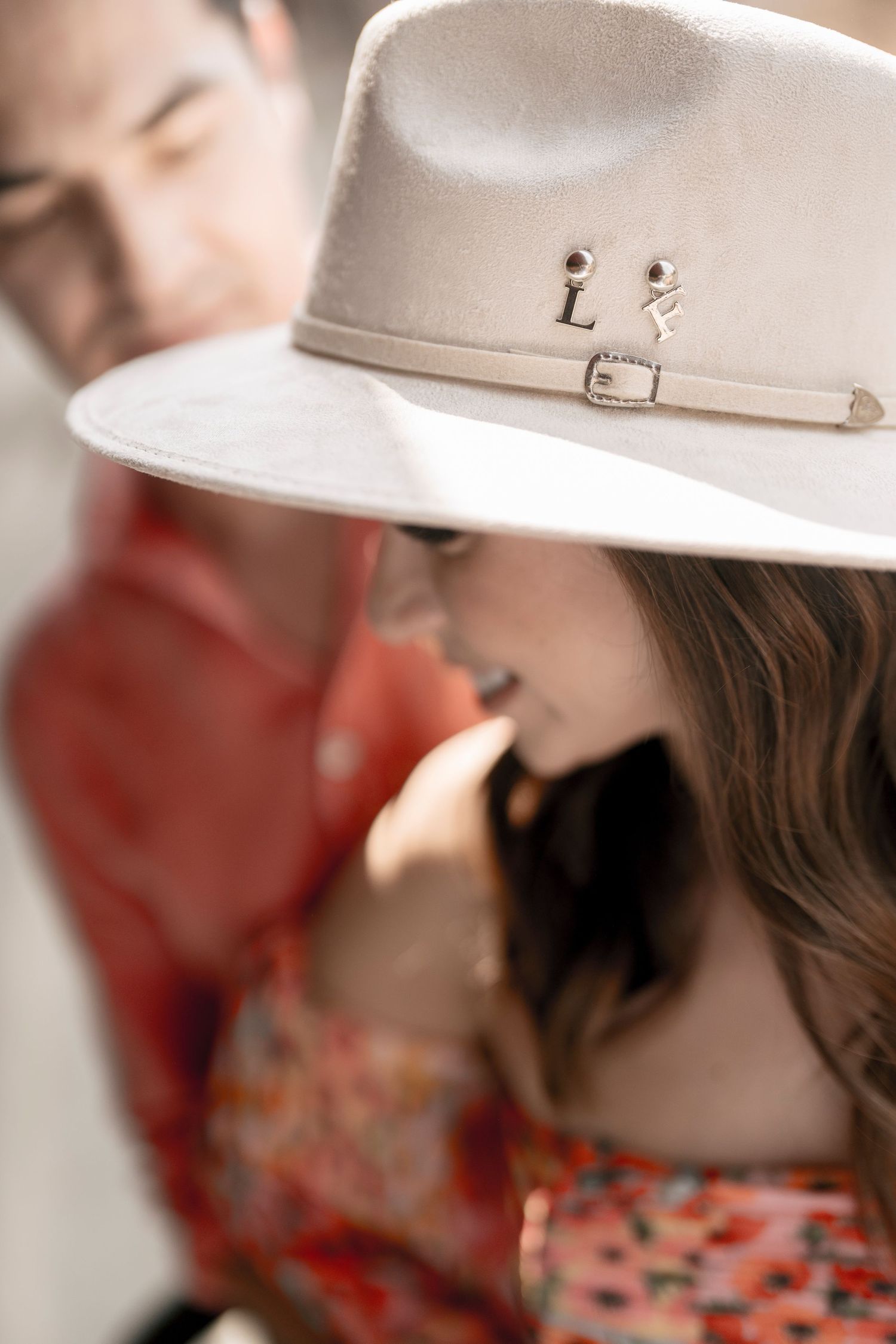 A woman in a wide-brim cream hat with pins smiles beside a man in a red jacket in a romantic outdoor setting.