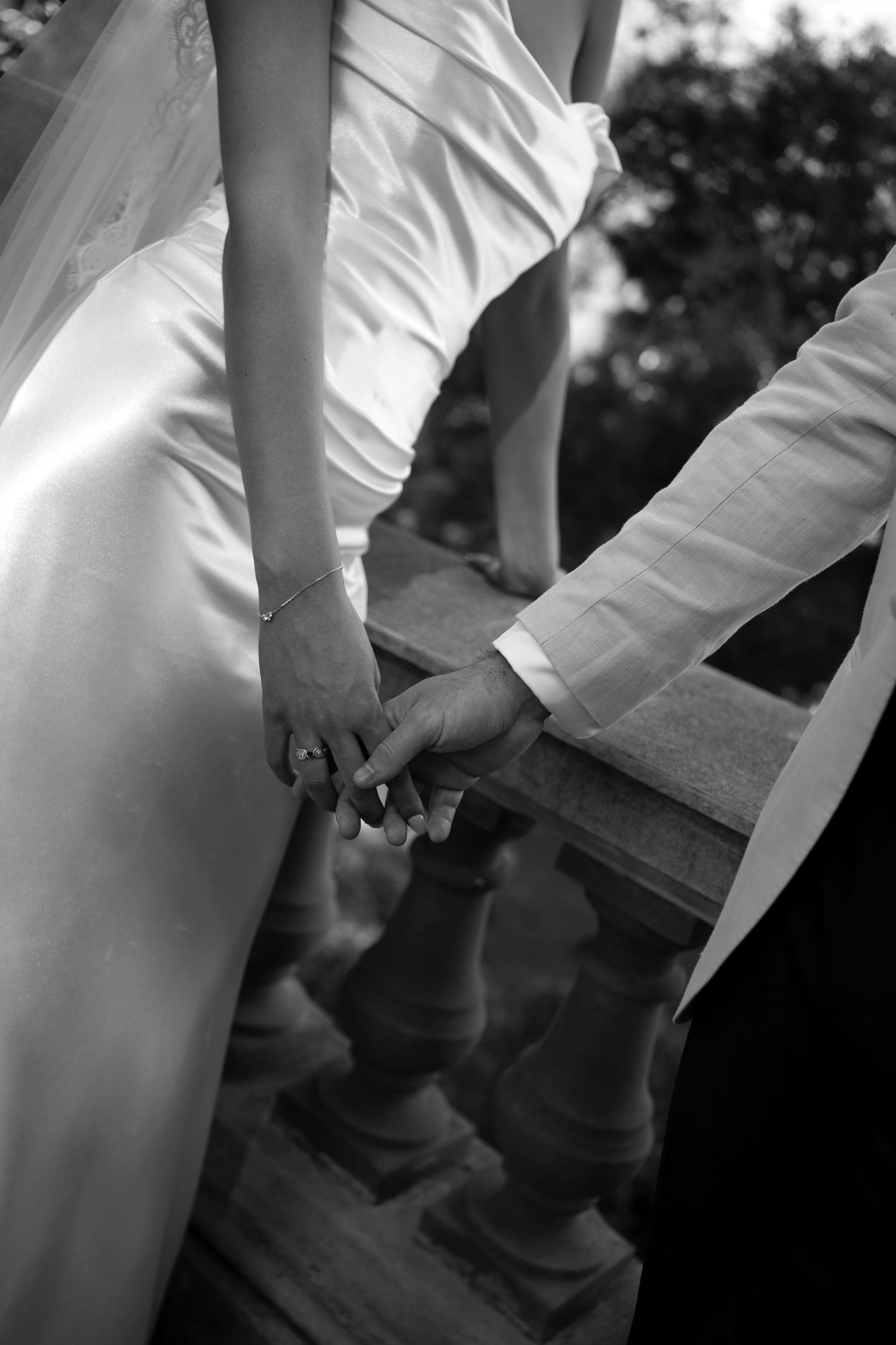 Black and white photo of bride and groom holding hands on elegant staircase, wedding rings visible.