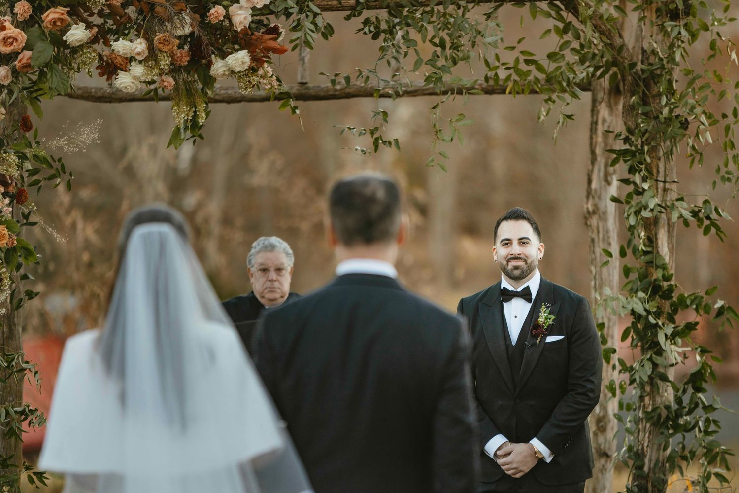Groom smiling at bride approaching outdoor wedding altar decorated with flowers and greenery.