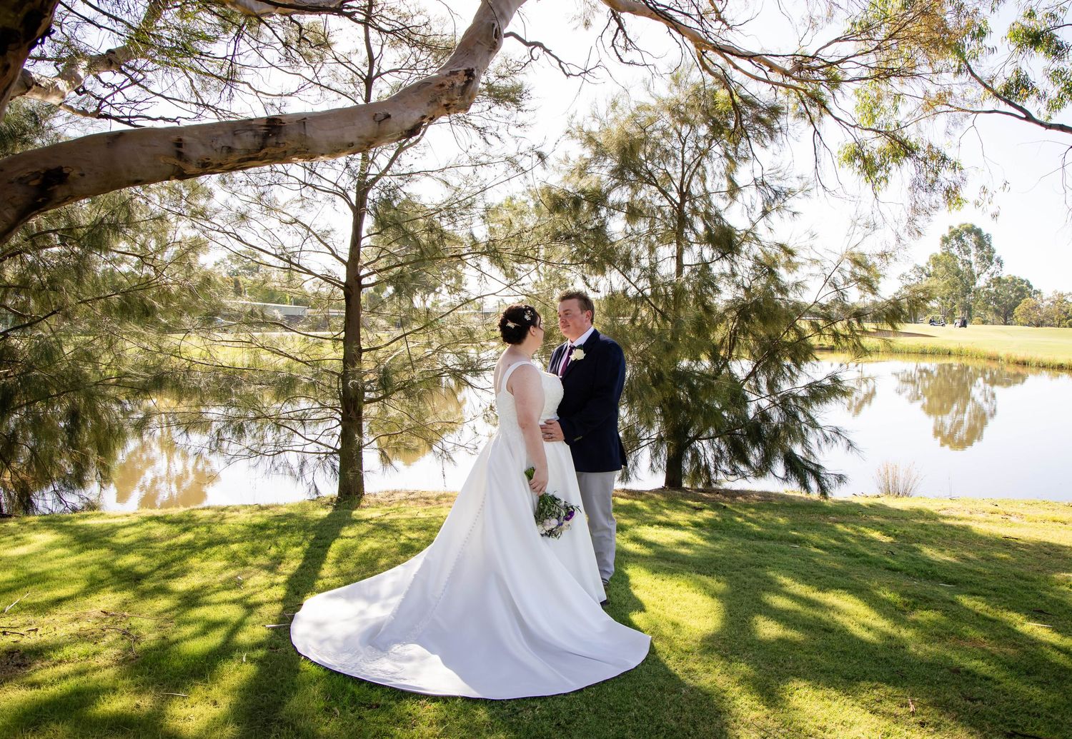 Bride and groom standing under a tree by a lake on their wedding day, bride wearing a long white gown.