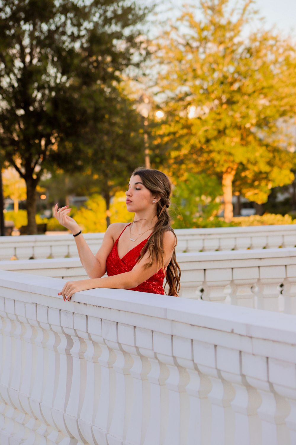 A young woman in a red sequin dress gazes thoughtfully while leaning on a white balustrade at sunset.