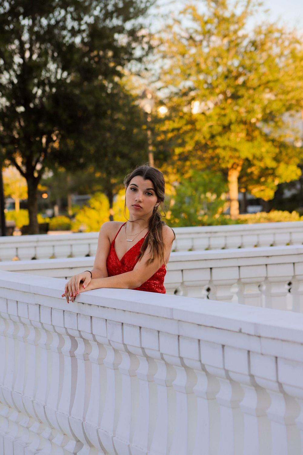 An elegant young woman in a red sequin dress leans against a white balustrade in a park at sunset.