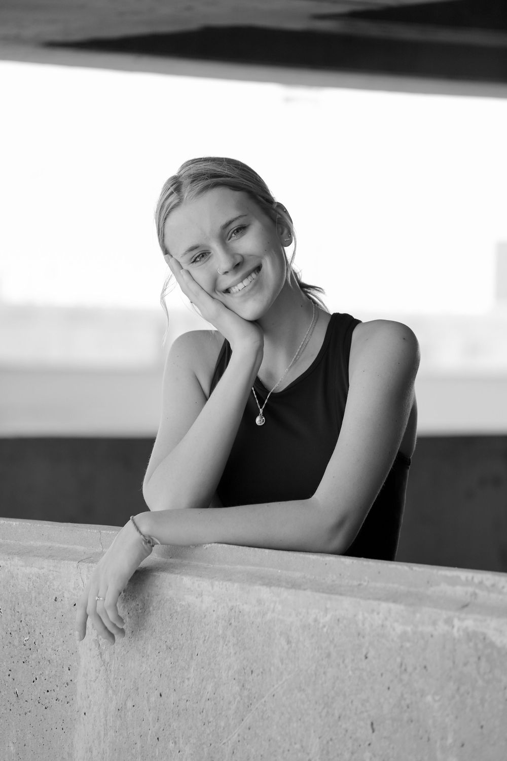 Black and white portrait of smiling young blonde woman leaning on concrete wall in black tank top.