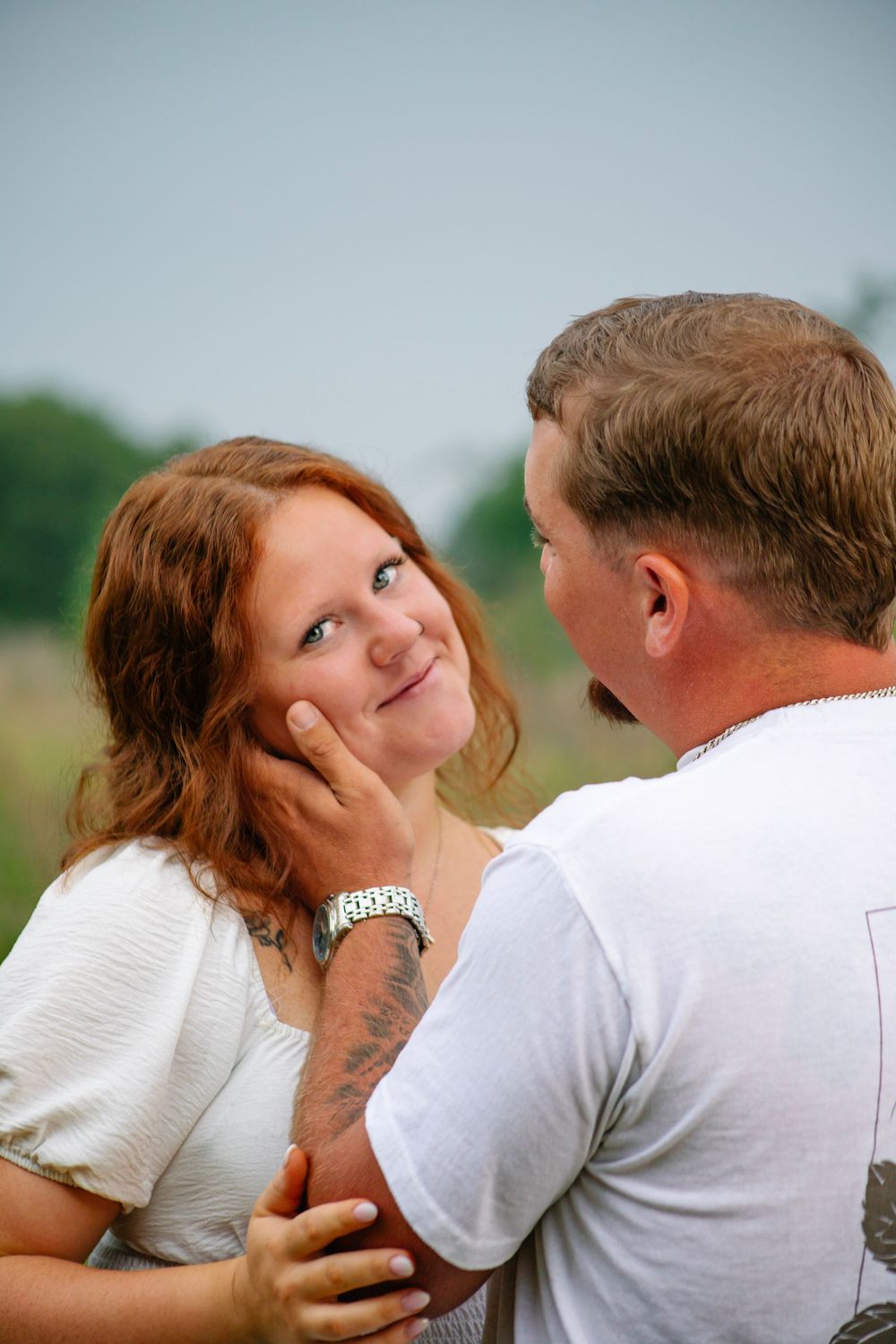 A man tenderly cups a redheaded woman's face as she smiles up at him in an outdoor field setting.