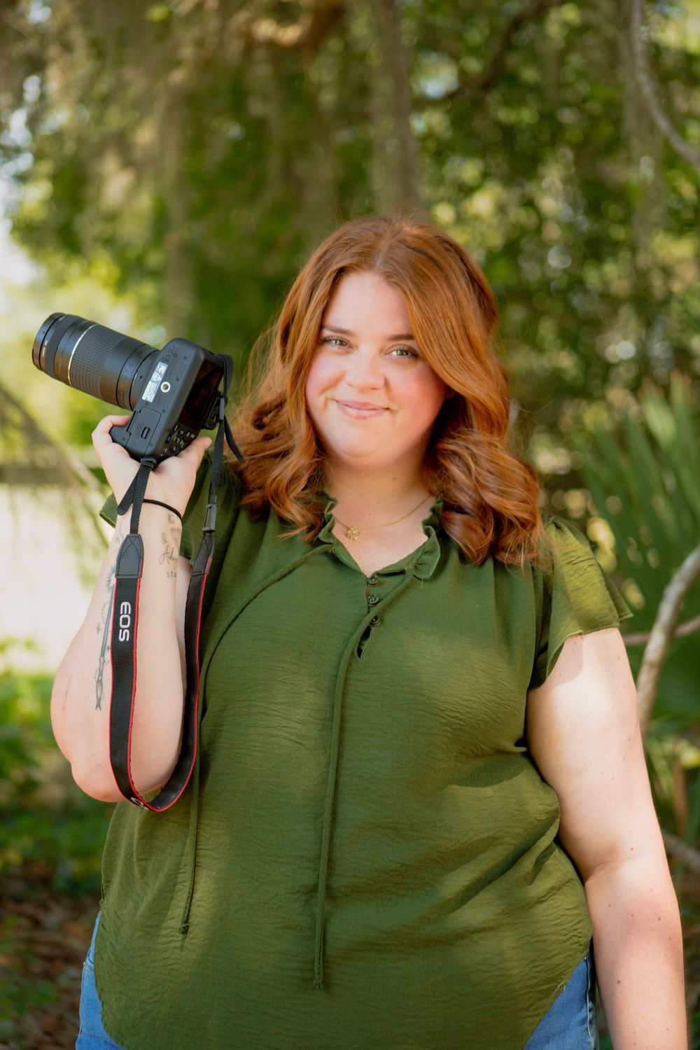 A young woman with red hair holds a DSLR camera outdoors, wearing a green top, surrounded by lush greenery.