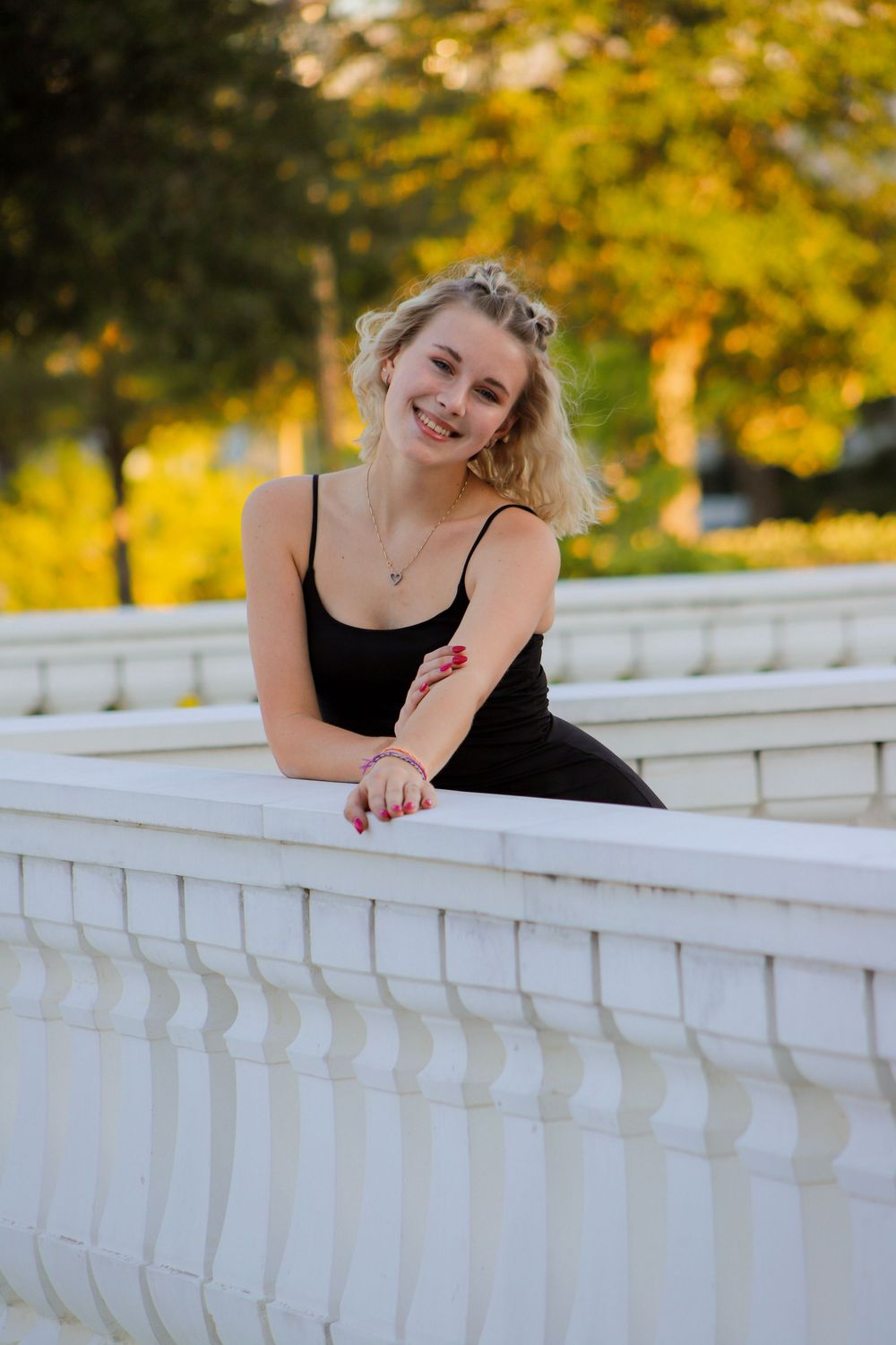 Smiling blonde woman in black dress leaning on white balustrade in sunny park.