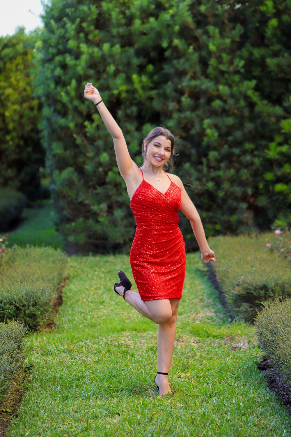 Young woman in red sequin dress posing ballet style in lush green garden.