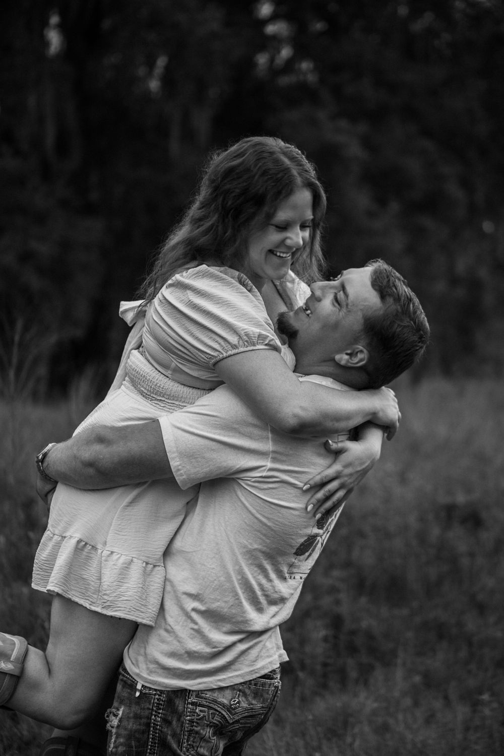 Black and white photo of a couple embracing outdoors, man lifting woman as she laughs joyfully in a field.