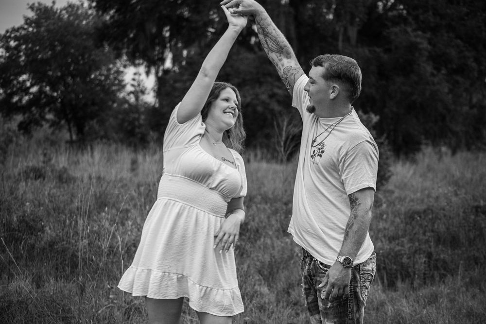 Black and white photo of a couple dancing outdoors in a field, man spinning woman in white dress.
