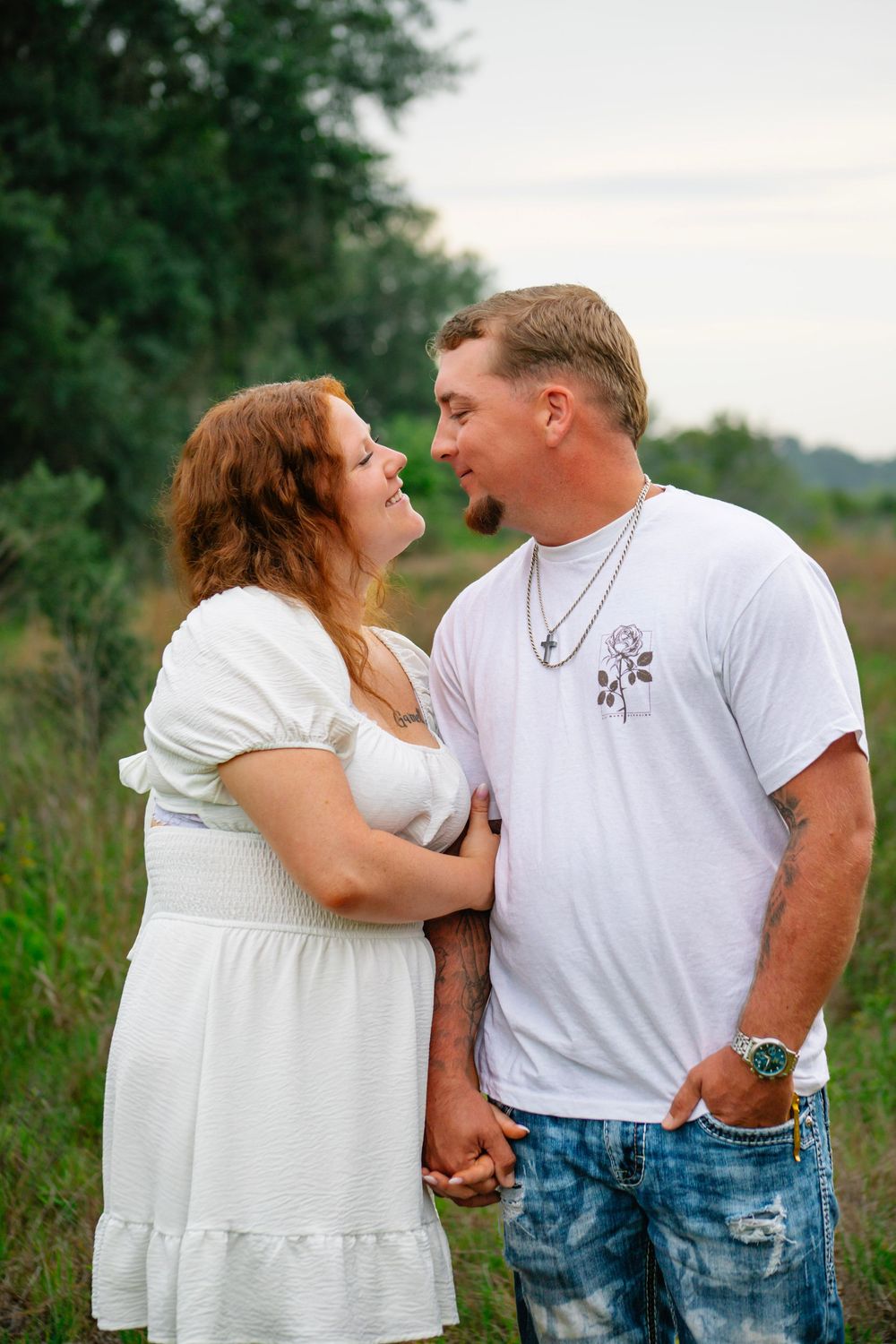 Couple holding hands and gazing at each other in a field, woman in white dress, man in white t-shirt and jeans.