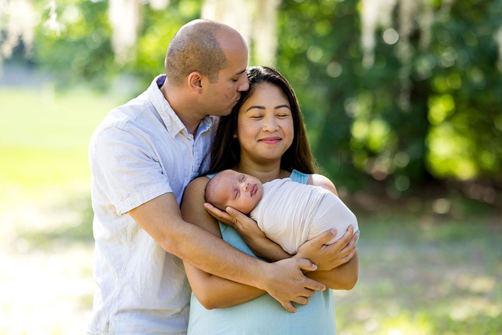 A couple tenderly holds their newborn baby outdoors, surrounded by lush green trees in a family portrait.