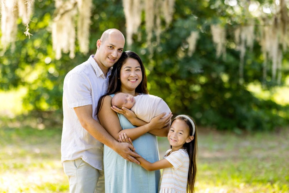Family of four poses outdoors, parents holding newborn baby with young daughter smiling beside them in a lush green park.
