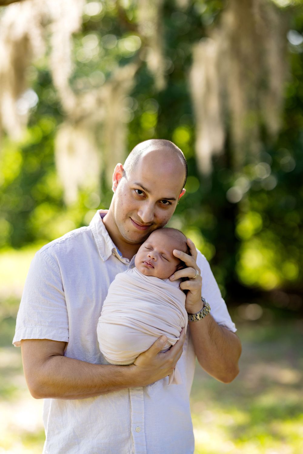 Bald father smiling and holding swaddled newborn baby outdoors in a lush green park setting.