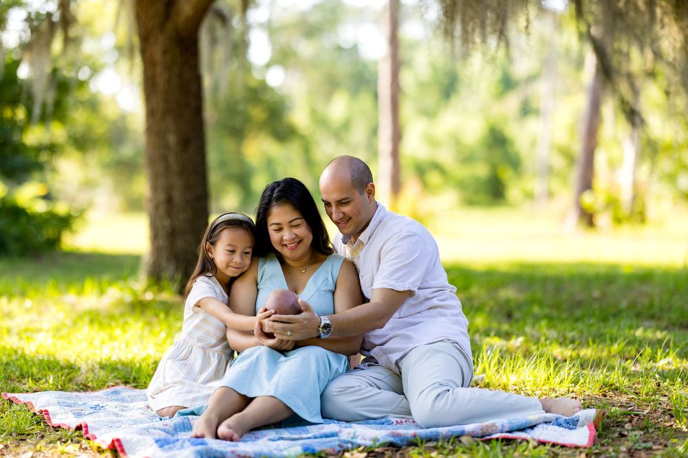 Family of four sitting on a blanket in a sunny park, parents holding newborn baby with young daughter beside them.