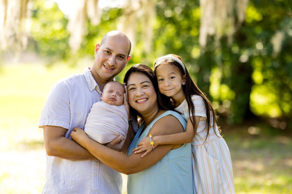 Happy family of four posing outdoors, parents holding newborn baby and young daughter, surrounded by lush green trees.