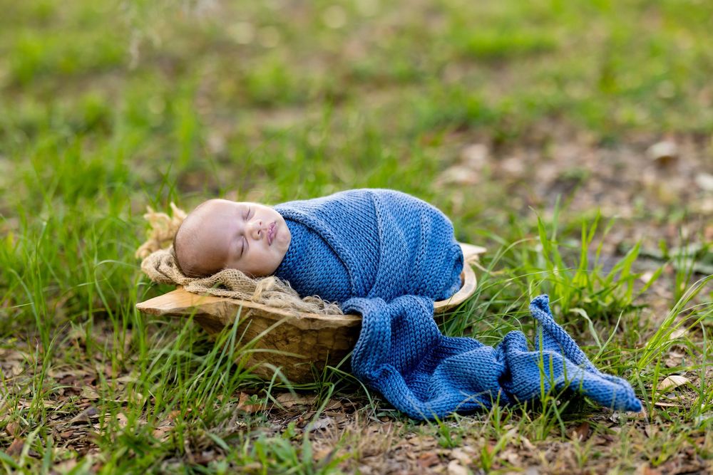 Newborn baby swaddled in blue knit wrap sleeping peacefully in a wooden bowl on green grass outdoors.
