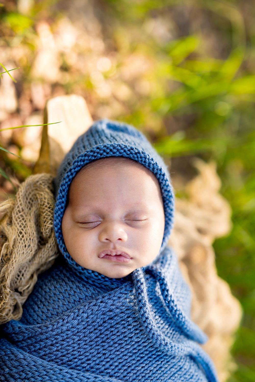 Sleeping newborn baby wrapped in blue knit swaddle and matching bonnet hat, outdoors in natural setting.