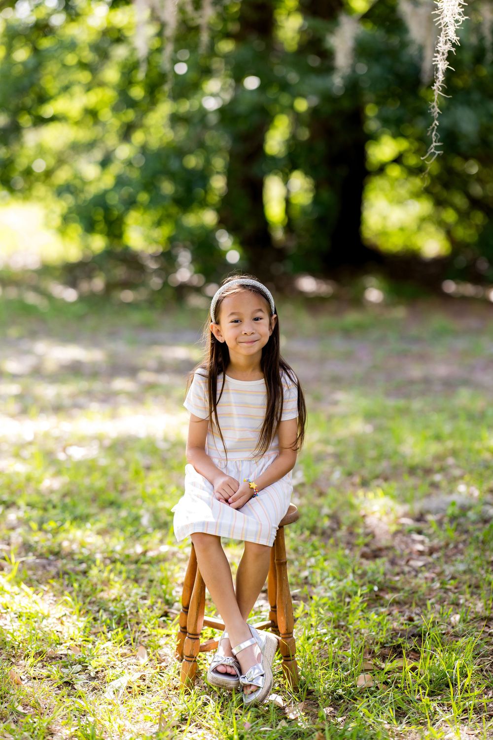 Young girl in striped dress sits on wooden stool outdoors, smiling at camera with lush green trees behind her.