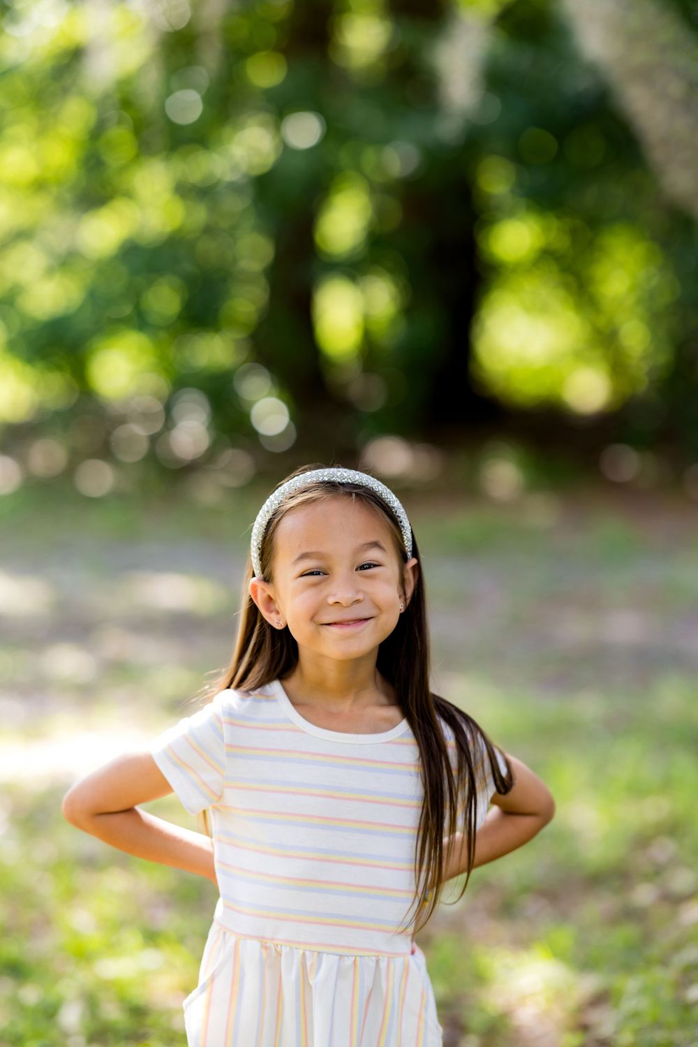 Smiling young girl with long hair wearing a striped dress and headband, posing outdoors in a sunny green park.