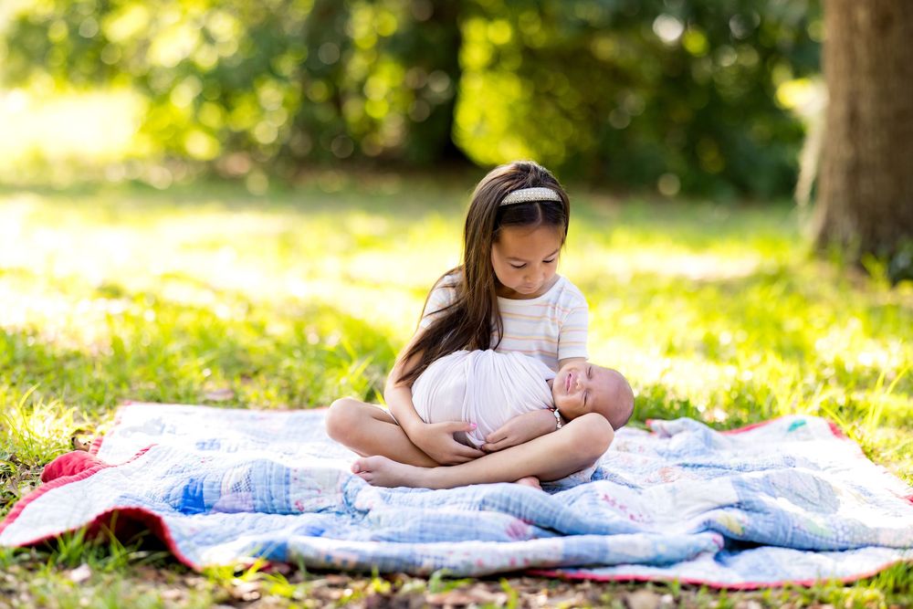 A young girl sits cross-legged on a quilt in a sunny park, gently cradling a newborn baby in her lap.