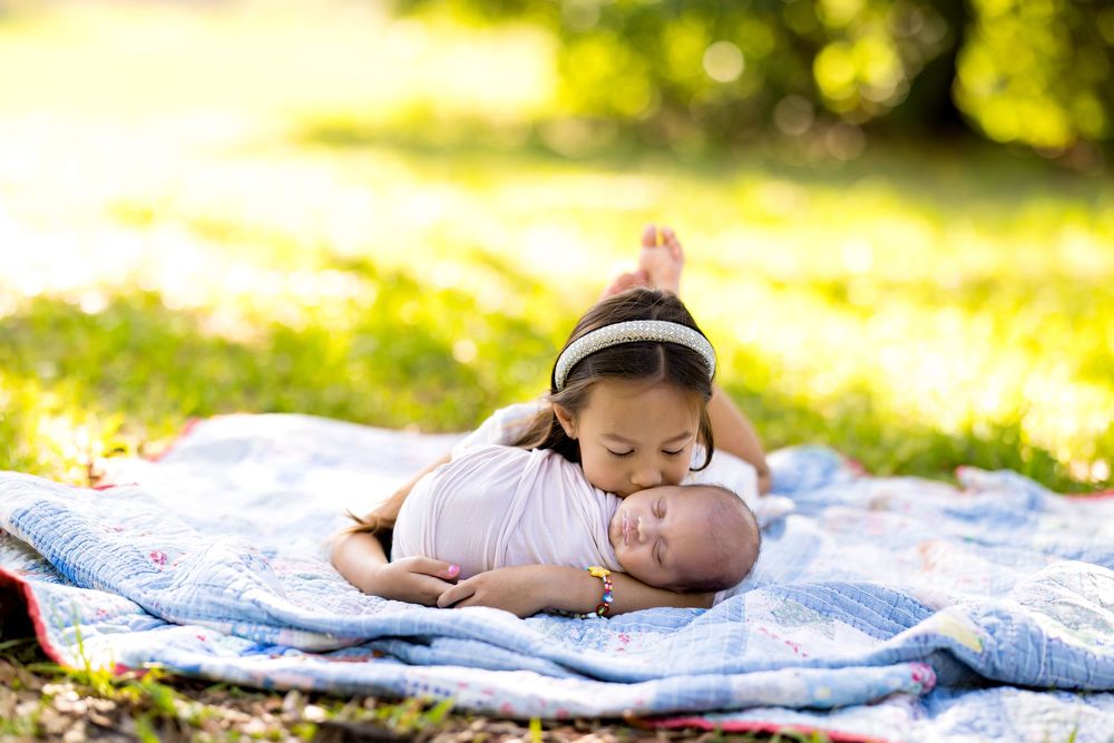 A young girl tenderly kisses a sleeping newborn baby on a colorful quilt outdoors in sunny green park.