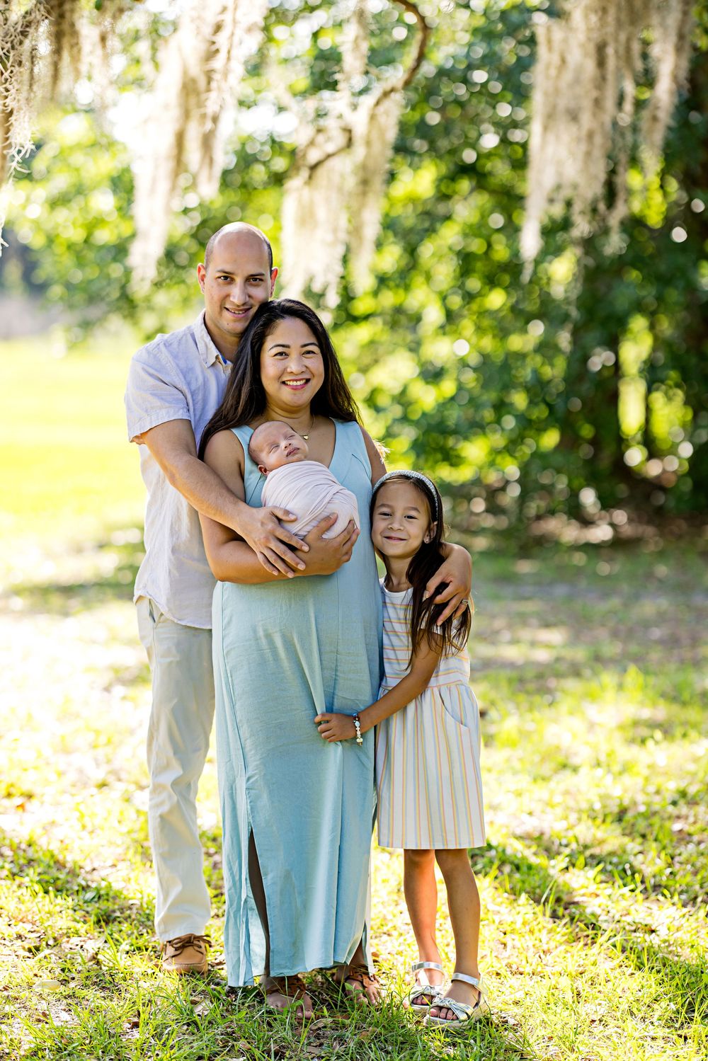 Family of four poses outdoors under Spanish moss trees, parents holding newborn baby with young daughter smiling beside them.