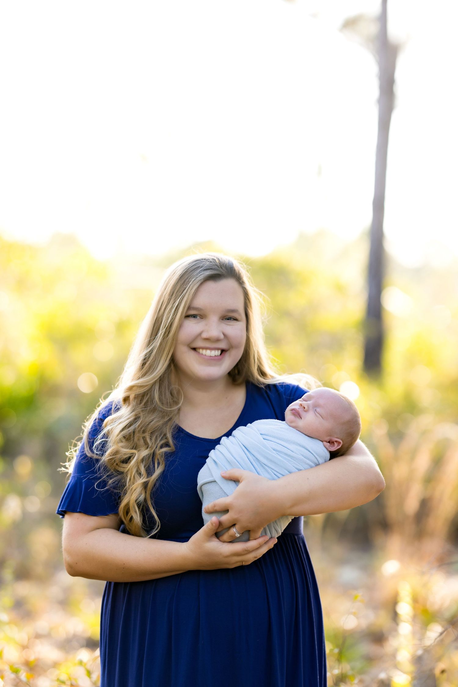 Young woman in navy dress smiling outdoors while cradling a swaddled newborn baby in a sunlit natural setting.