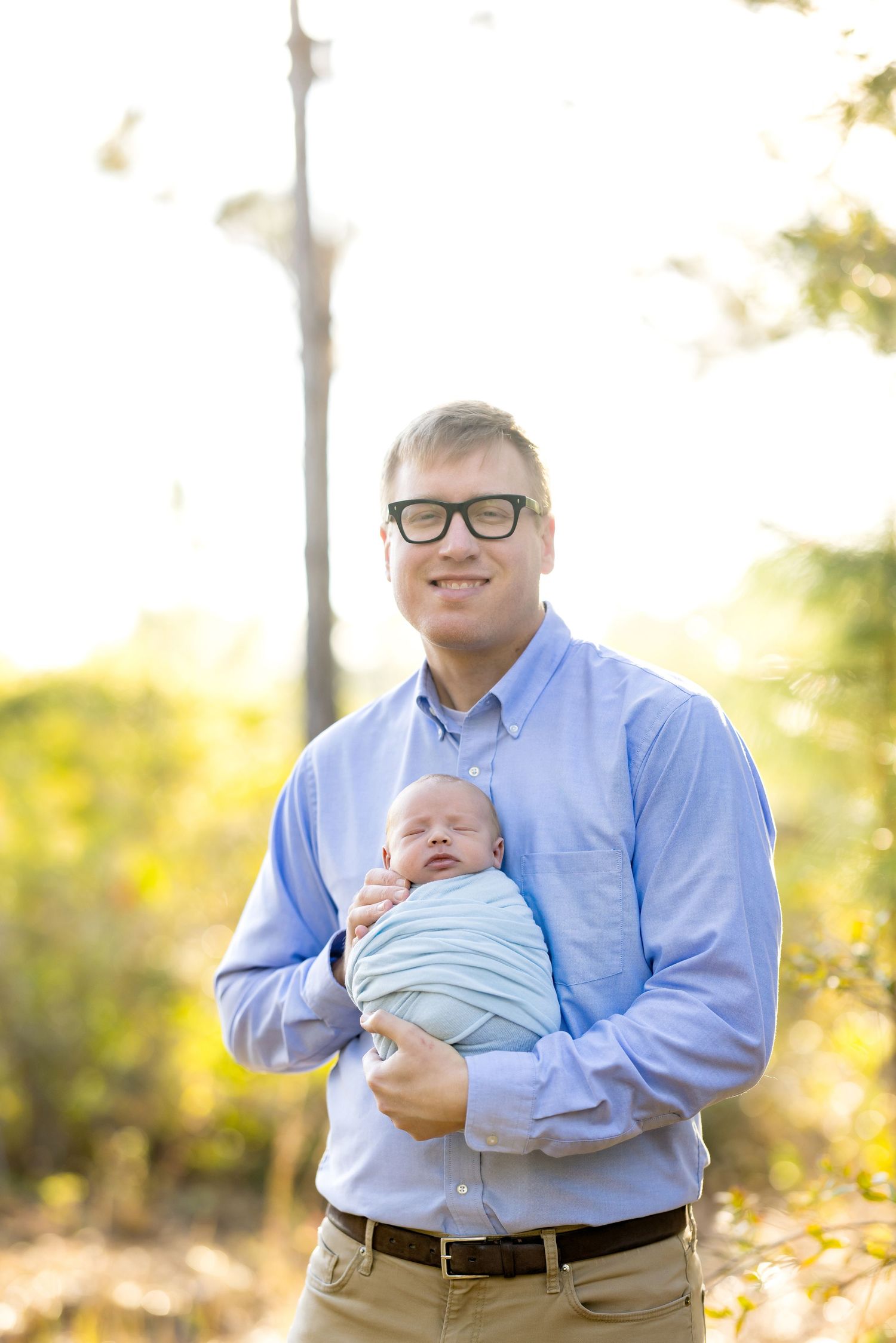 Smiling man with glasses holding a sleeping swaddled newborn baby outdoors in a sunny green park setting.