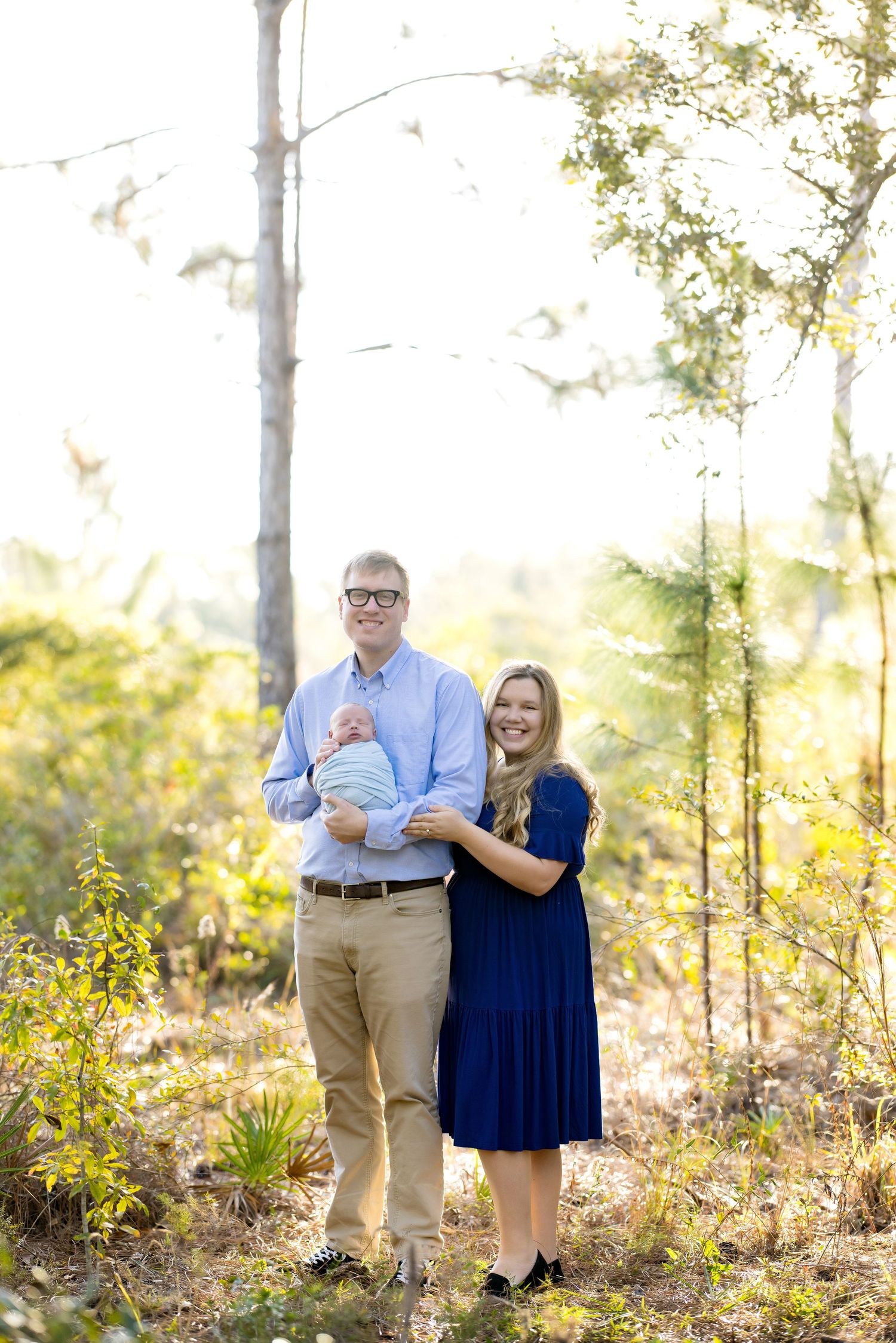 A smiling couple stands outdoors in a sunlit forest, holding their newborn baby, dressed in blue and navy attire.