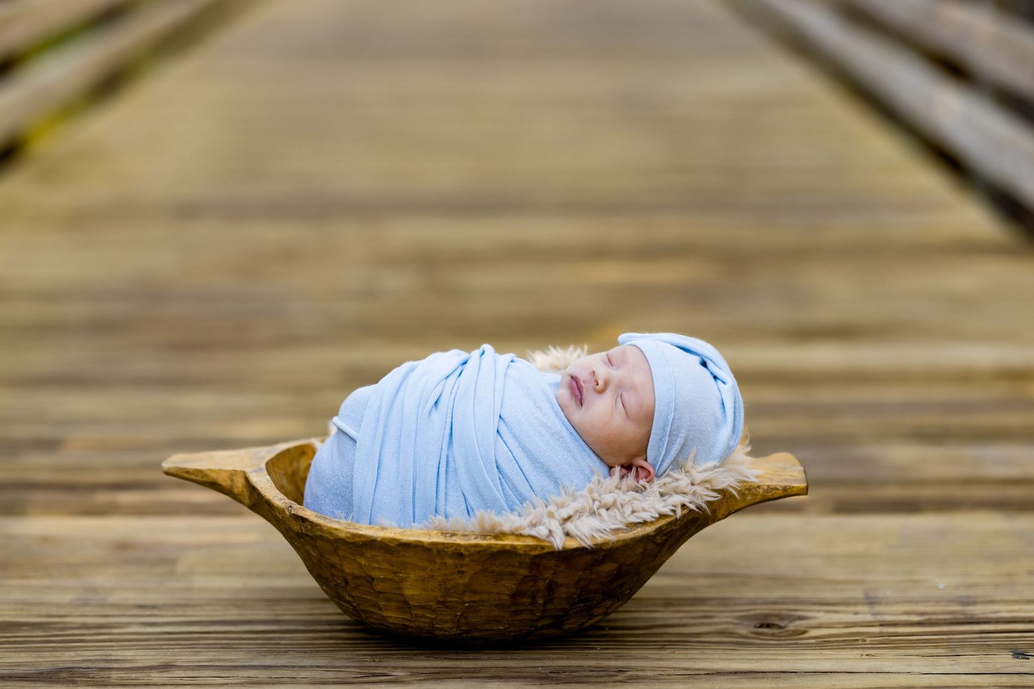 Newborn baby wrapped in blue swaddle sleeping peacefully in a wooden bowl on a rustic bridge.