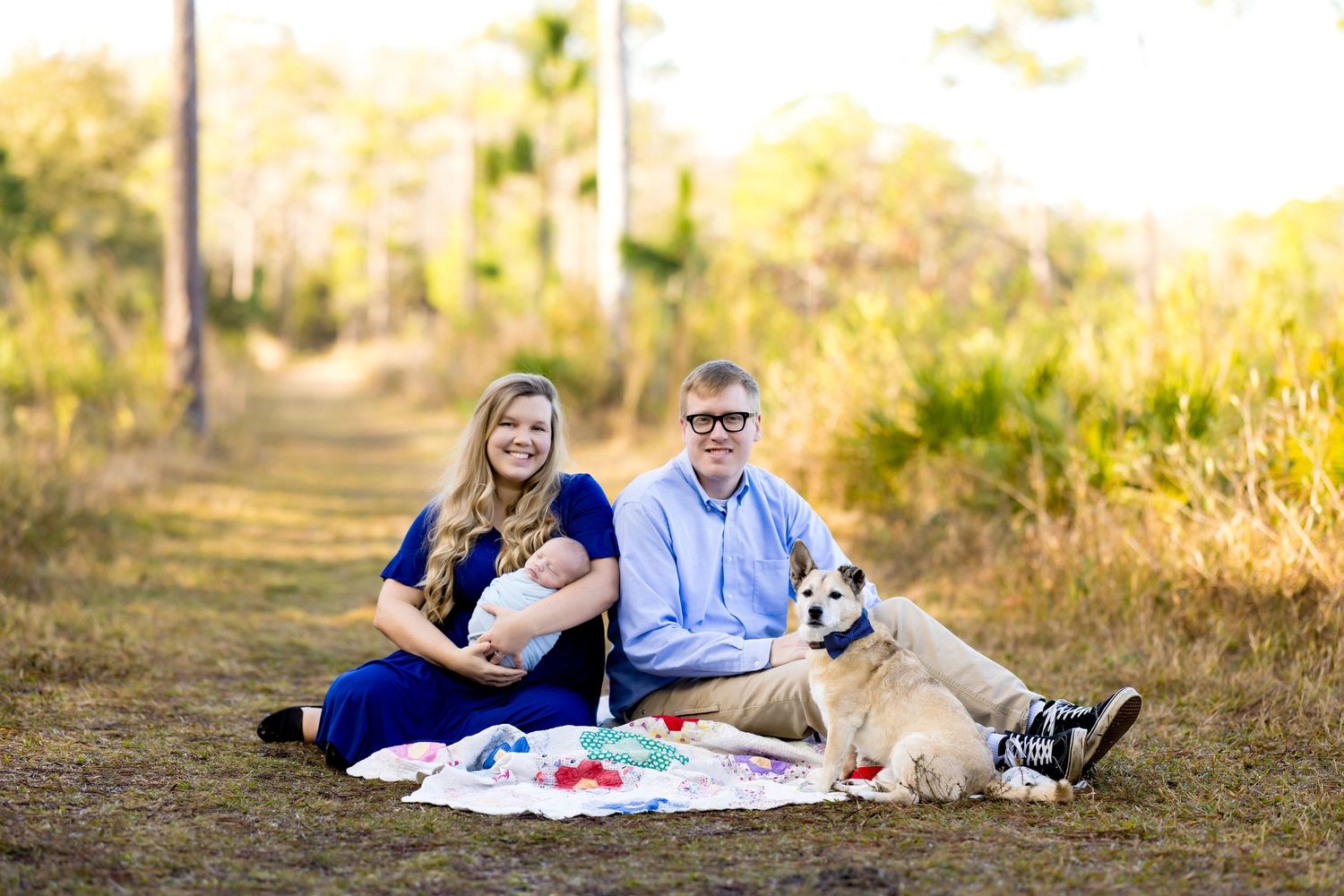 Family with baby and dog enjoying a picnic outdoors on a sunny day in a scenic park setting.