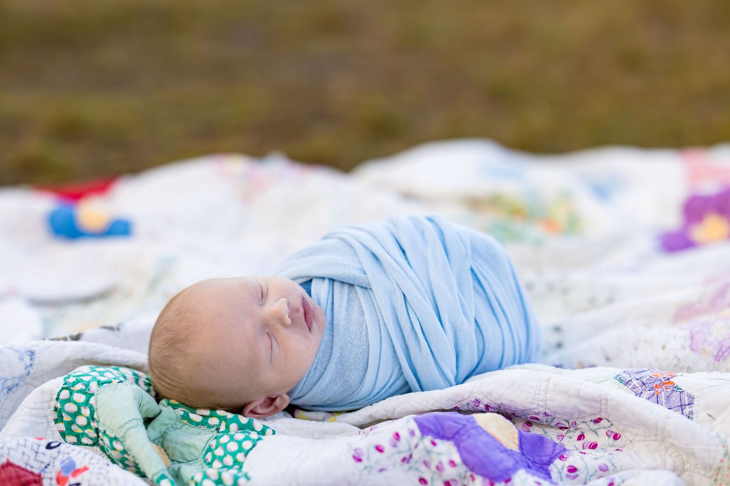 Newborn baby swaddled in light blue wrap sleeping peacefully on a colorful quilt blanket outdoors.
