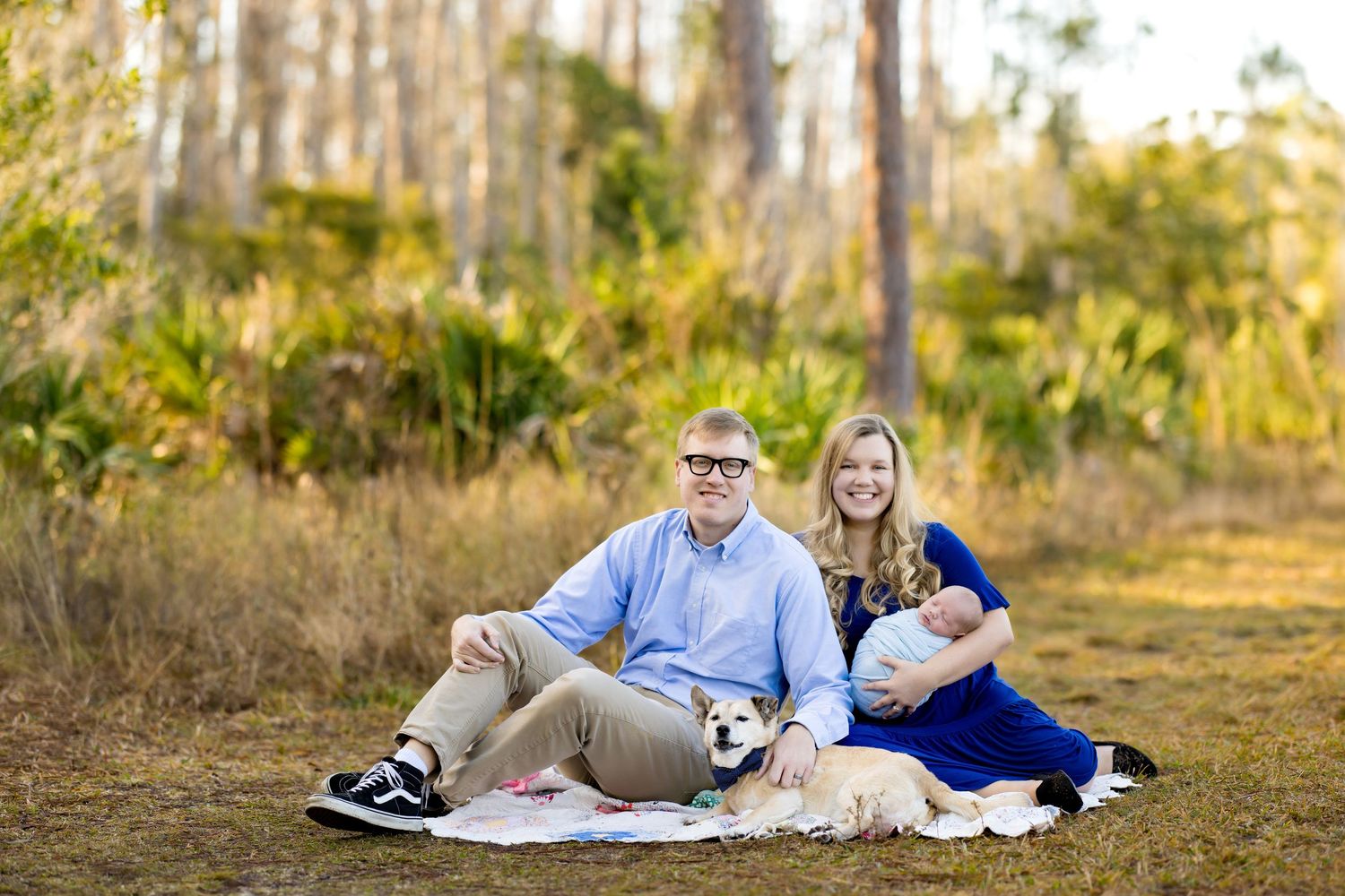 Family portrait with couple, newborn baby, and yellow lab dog sitting on blanket in sunny outdoor field.