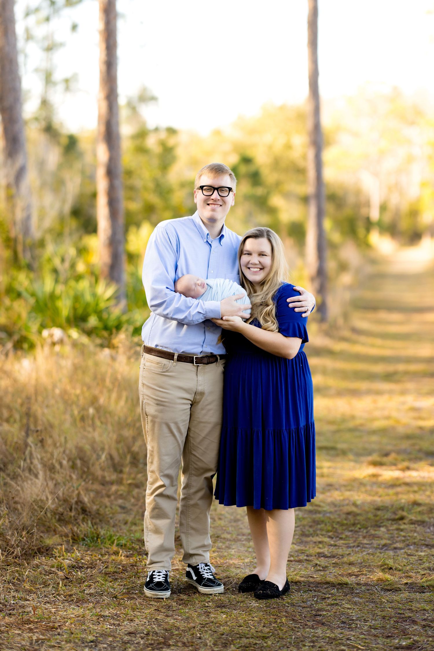 Couple poses outdoors in a pine forest path, man in blue shirt and woman in royal blue dress holding a newborn baby.