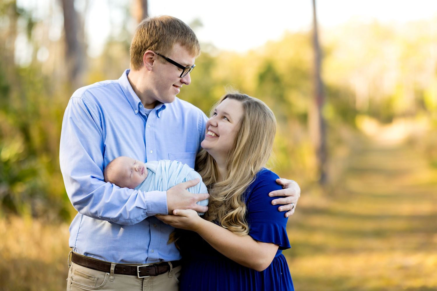 Young couple smiling at each other while holding their newborn baby outdoors in a sunlit park setting.
