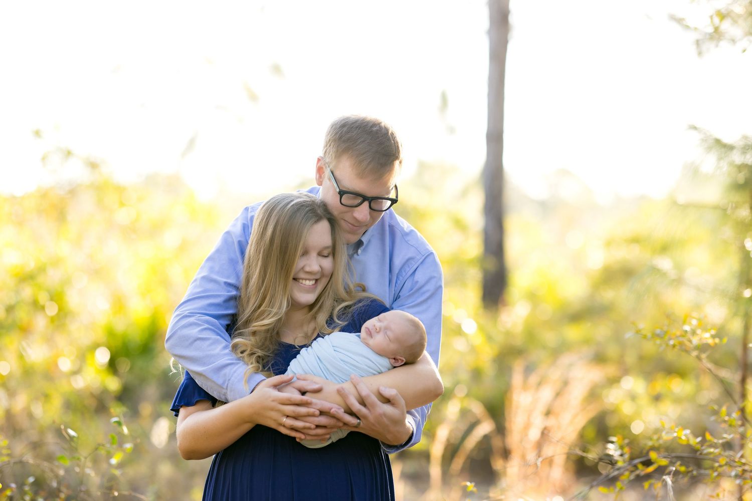 Happy couple smiling and cradling their newborn baby outdoors in a sunlit natural setting during a family portrait session.