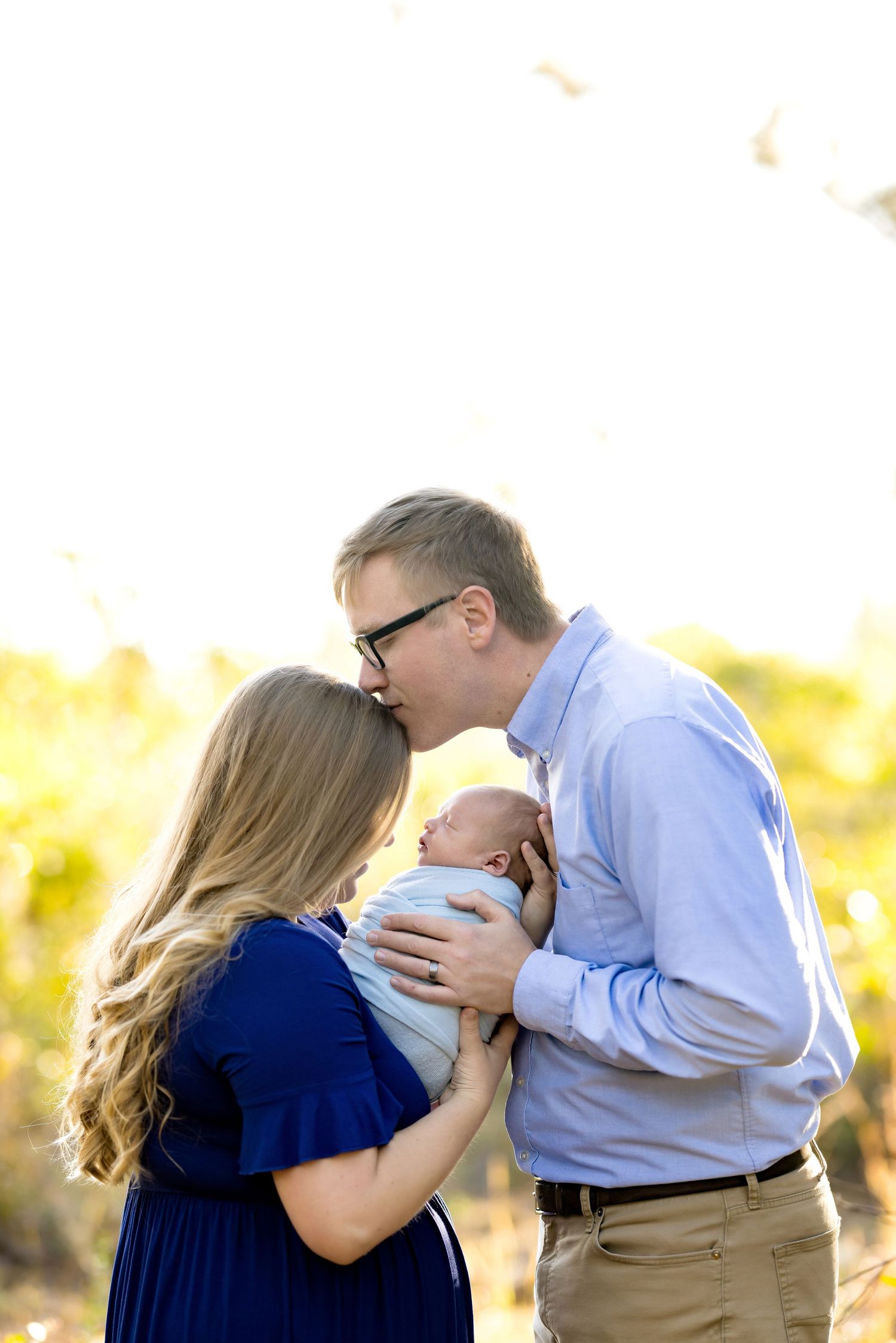 Parents share a tender kiss while cradling their newborn baby outdoors in golden sunlight during a family portrait session.