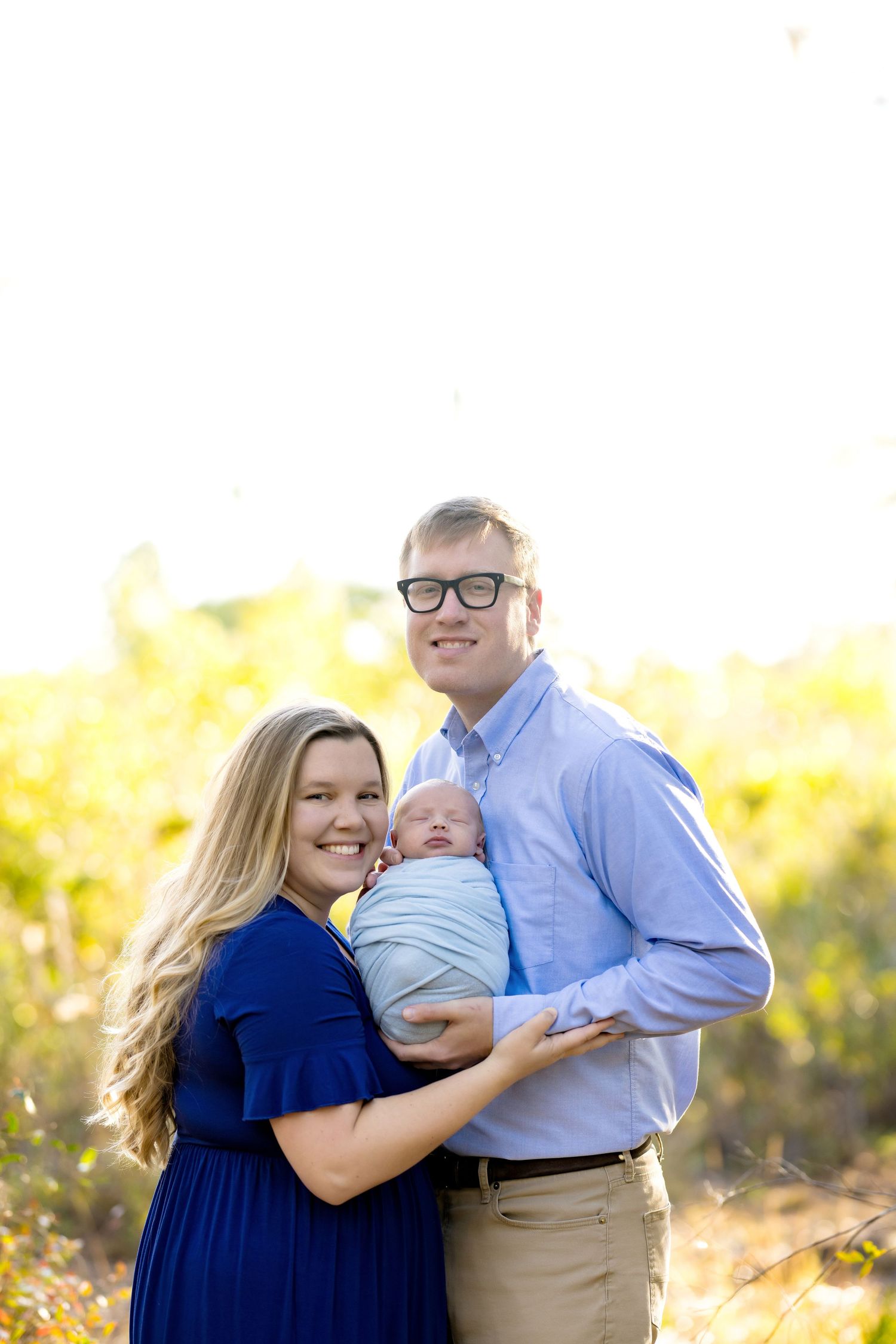 Young couple smiling outdoors holding swaddled newborn baby, woman in blue dress, man in light blue shirt, golden foliage background.
