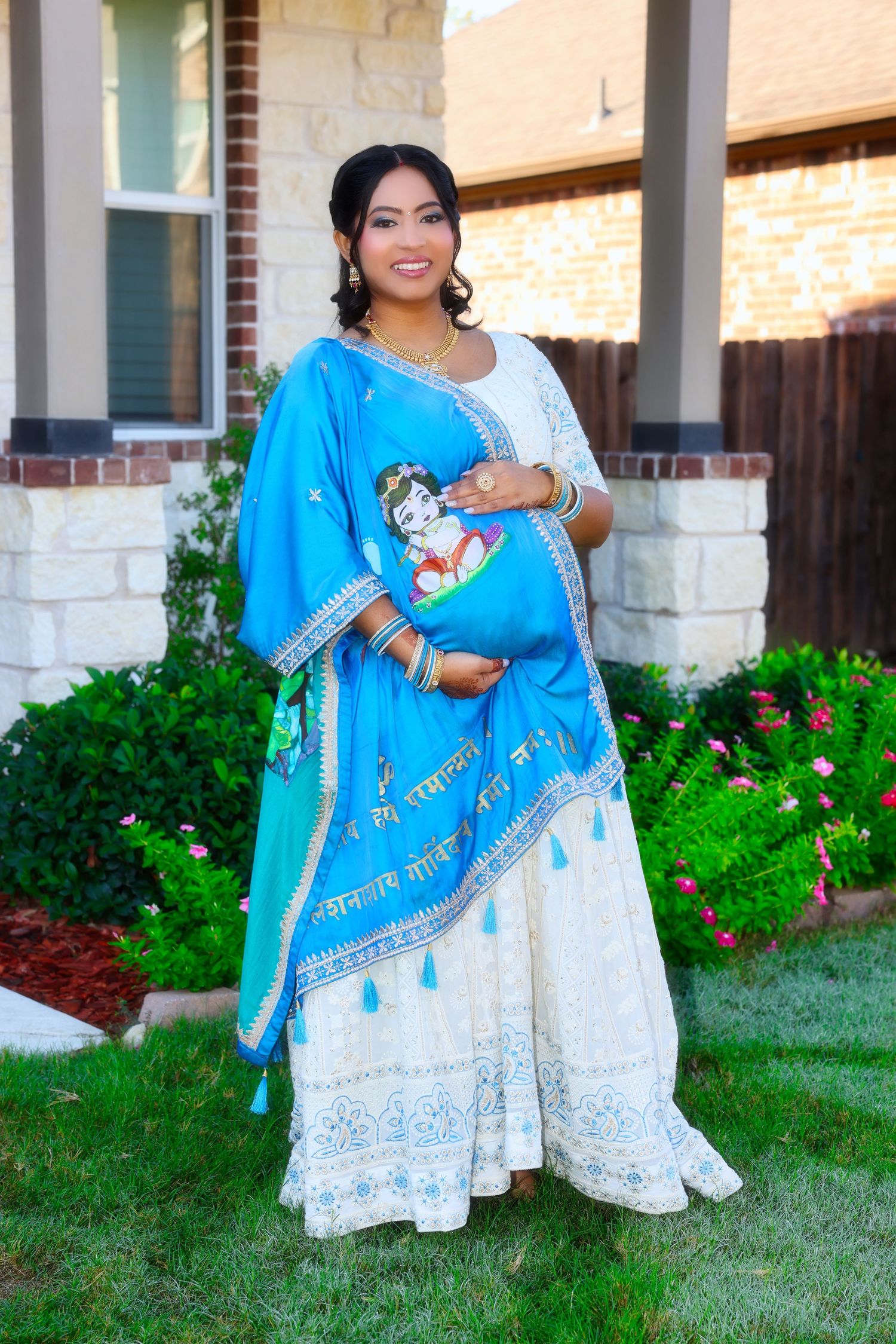 Pregnant woman in traditional blue and white Indian lehenga with dupatta, posing outdoors near brick home.