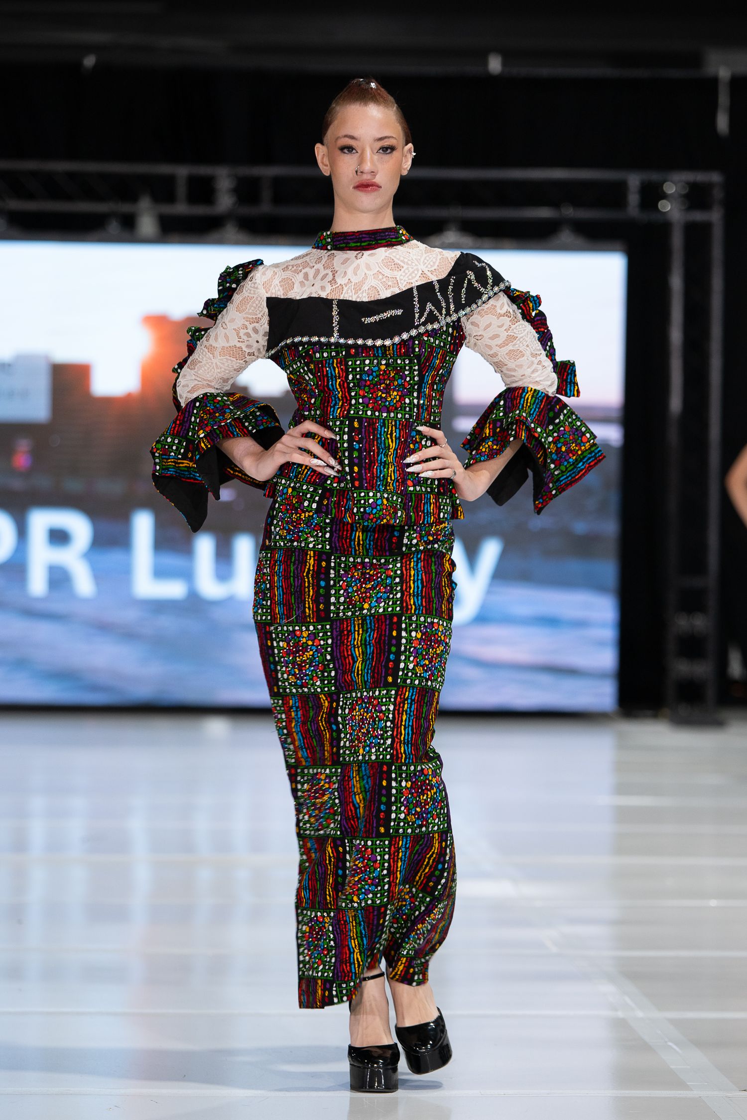 Model walks runway in colorful patterned gown with lace bodice and ruffled sleeves at fashion show.