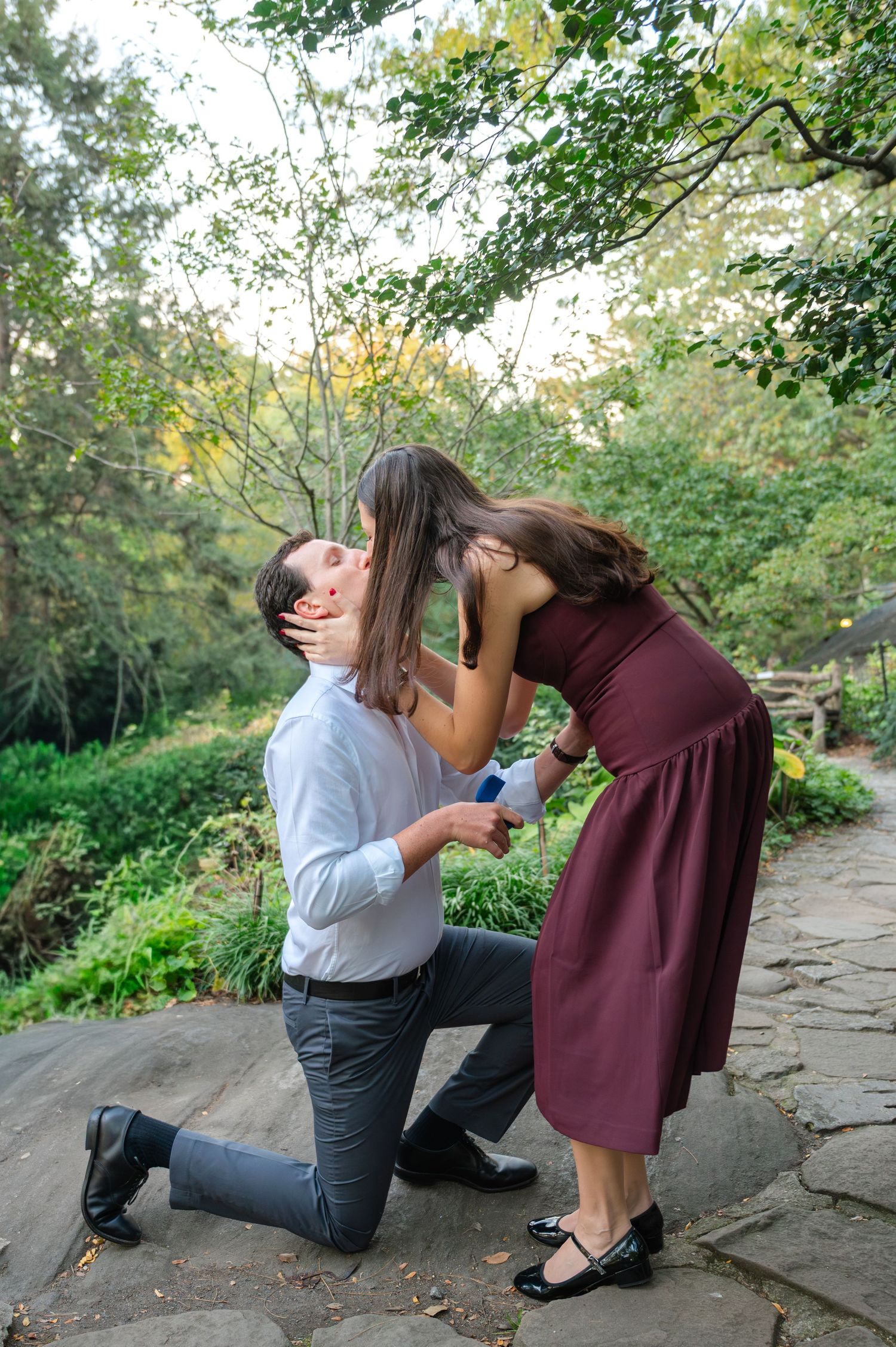 Man kneeling on one knee proposing to woman in burgundy dress in the Shakespeare Garden of Central Park on a warm October evening.