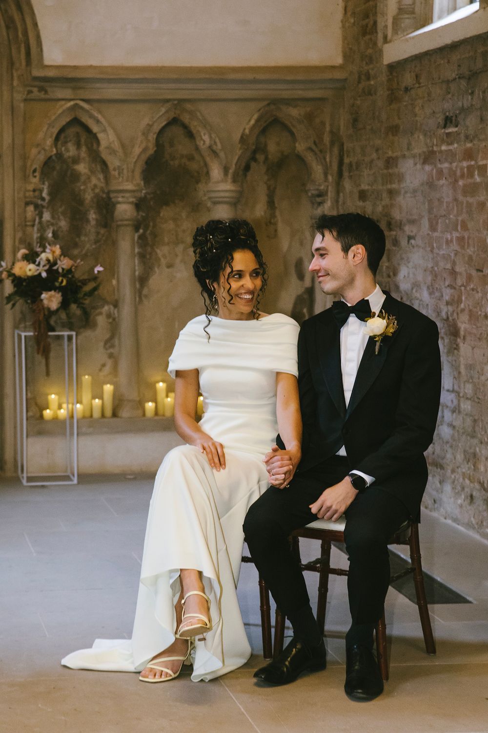Bride and groom sitting together in elegant gothic venue, bride in white gown, groom in black tuxedo with boutonniere.