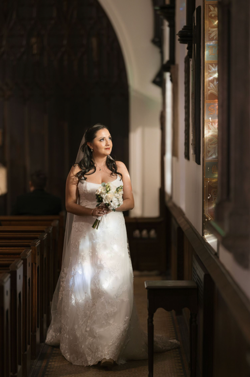 Bride in white lace gown holding bouquet stands in church aisle, bathed in natural light from stained glass window.