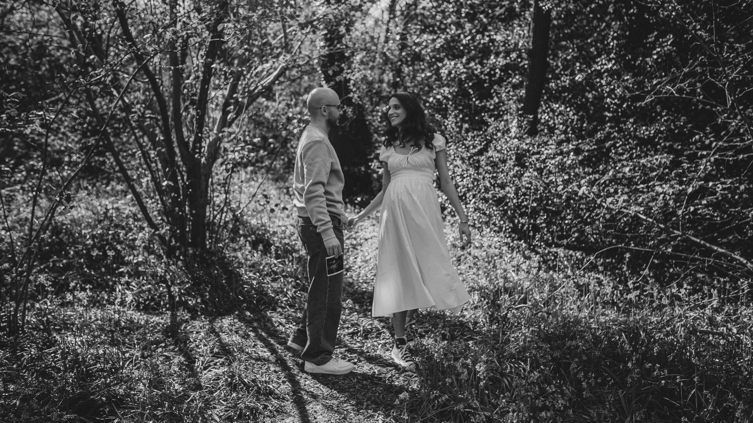 Black and white photo of a couple holding hands in a lush forest, bride in white dress.