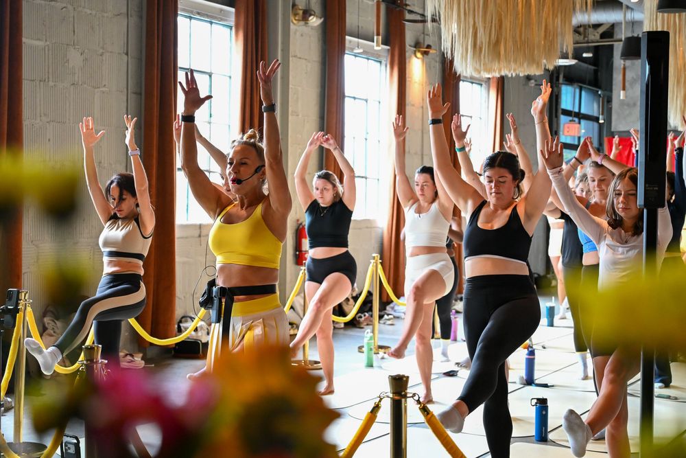 Group of women in activewear doing a yoga tree pose with arms raised in a bright studio fitness class.